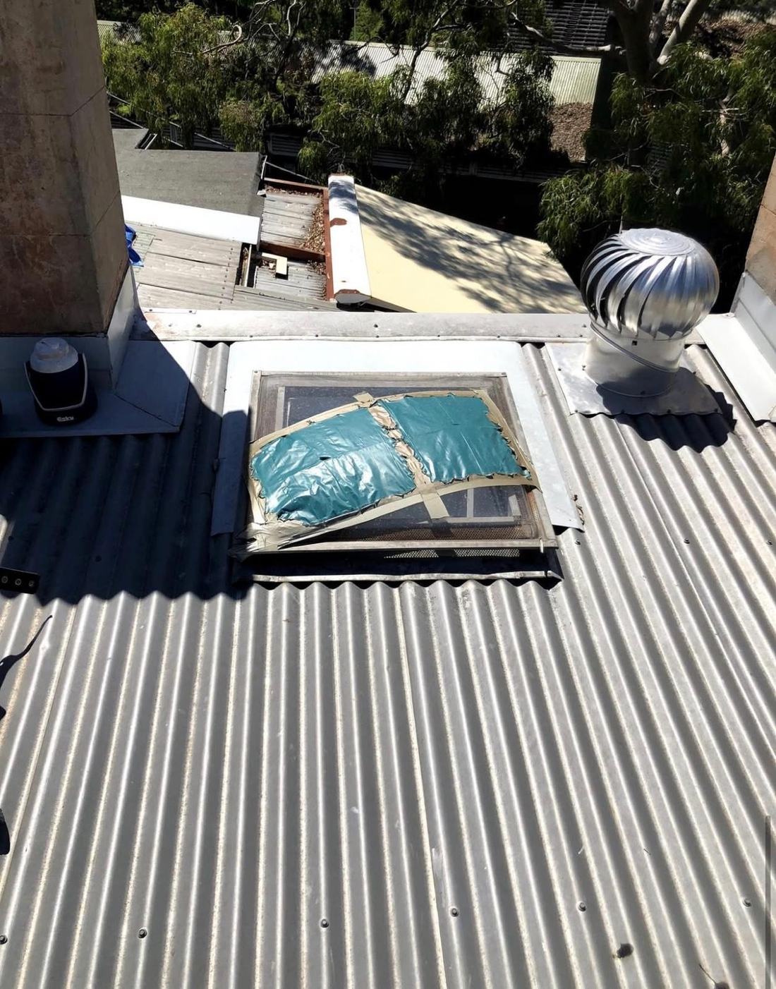 Broken skylight on a corrugated iron roof, patched over with a blue tarp and tape, beside a whirlybird vent