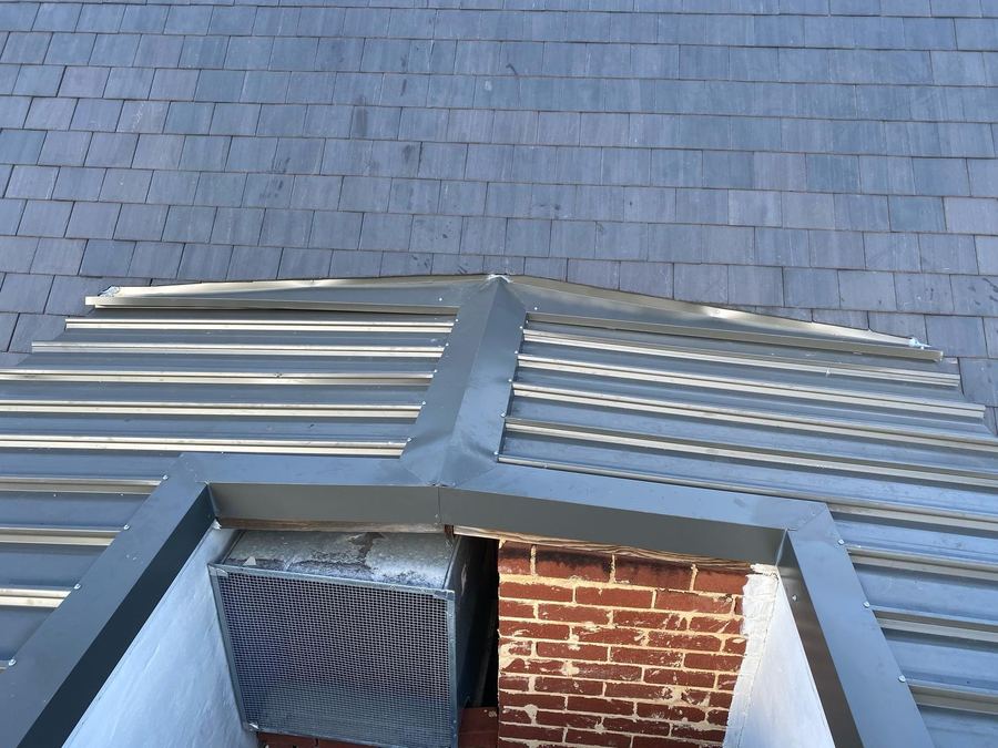 New grey metal tray roof with flashings wrapped around a brick chimney and a meshed vent, below a slate roof